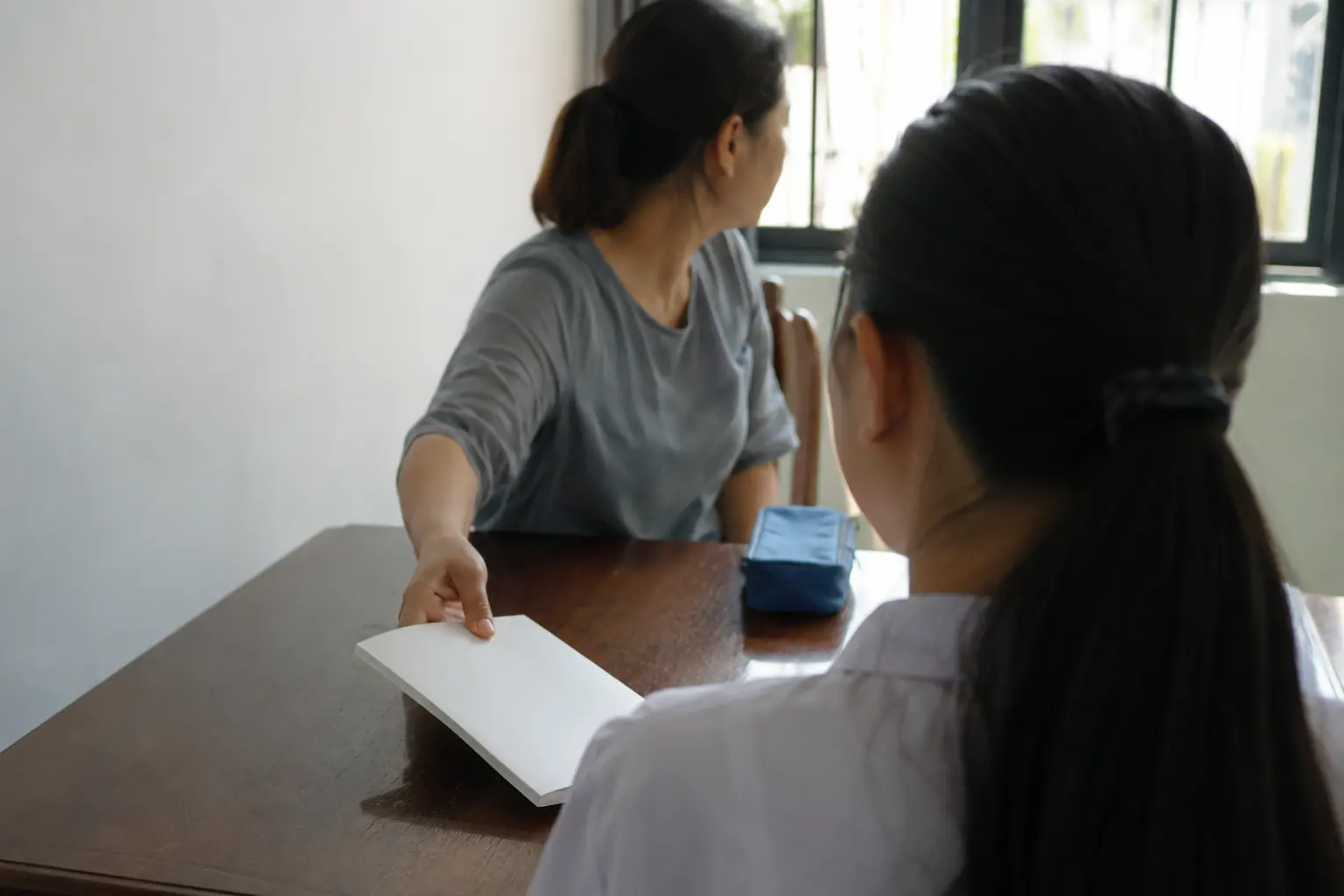 A tutor passing an exercise book across the table to a student, seen from behind the student.